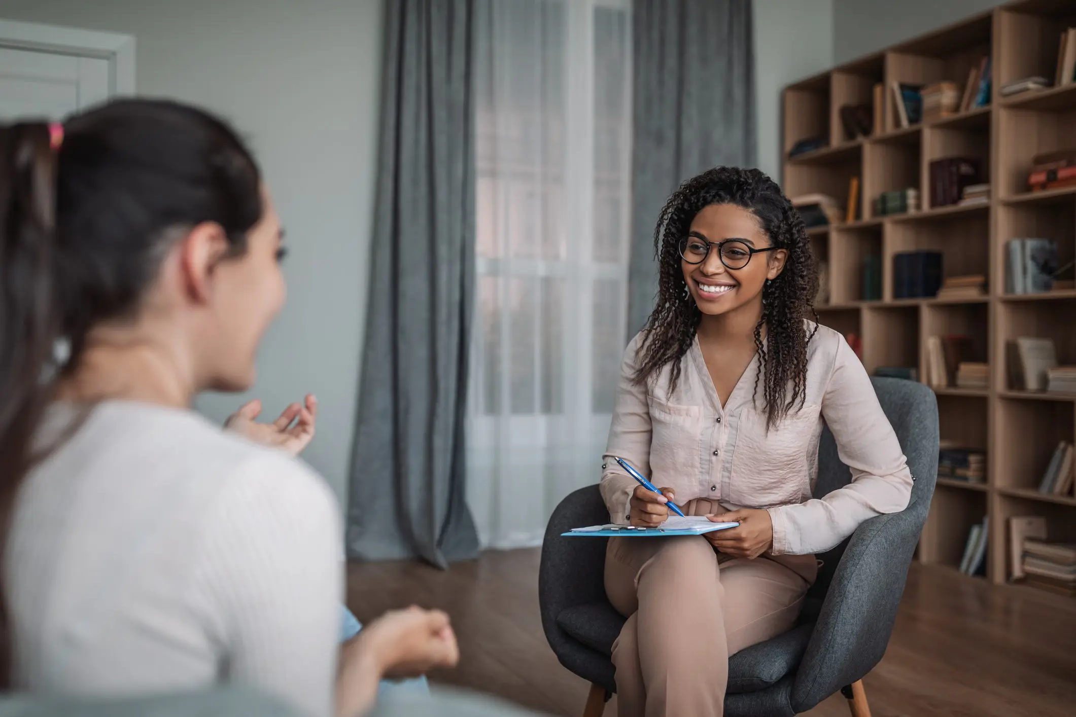 A smiling African American psychiatrist takes notes as she listens to her female patient, who sits across from her