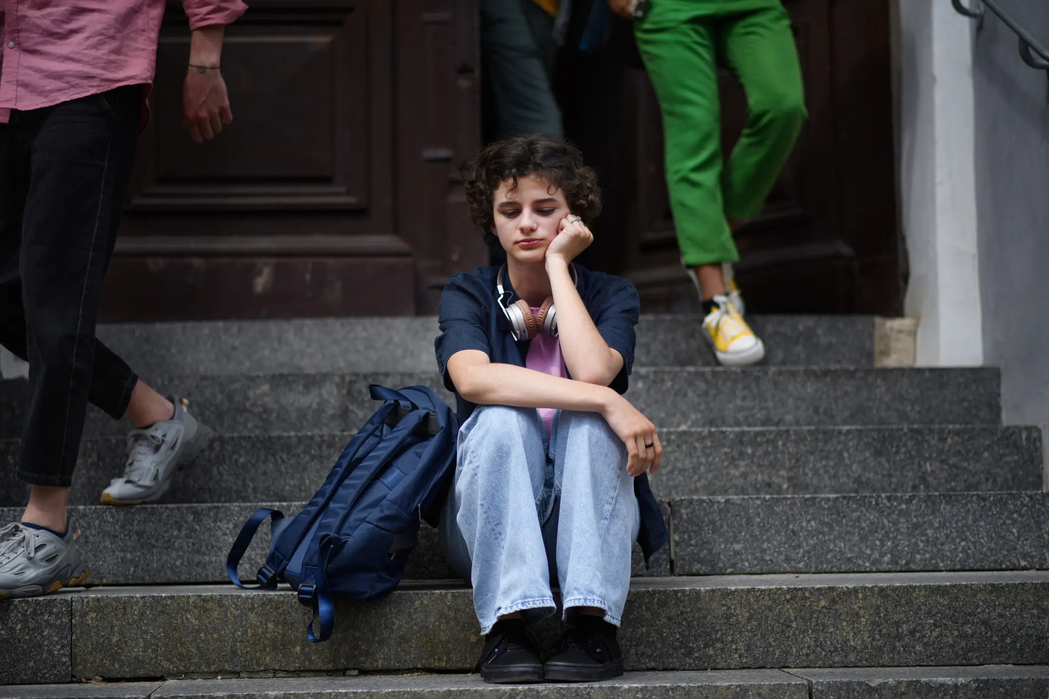 A teen boy sits on the steps in front of his school alone as others walk by him