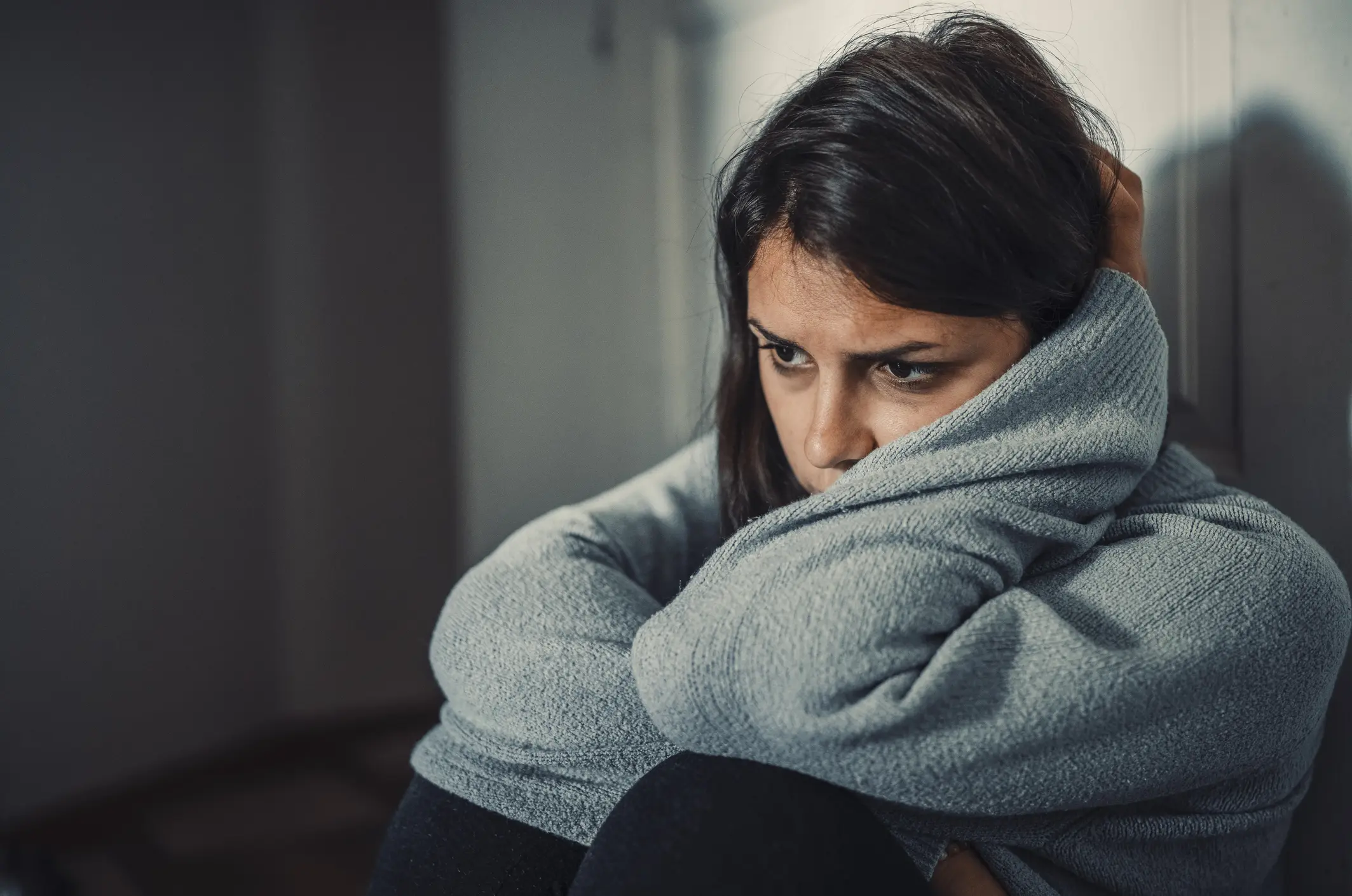 Distressed woman in a grey hoodie sits on the ground with her arms wrapped around herself.
